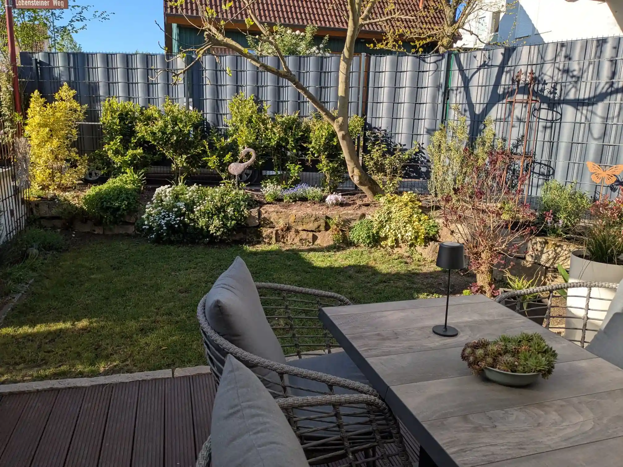 Nachher-Ansicht derselben Gartenfläche nach der Umgestaltung durch Molle GaLaBau: Moderne Holzterrasse, gemütliche Sitzgruppe, Natursteinmauer und blickdichter Sichtschutzzaun.