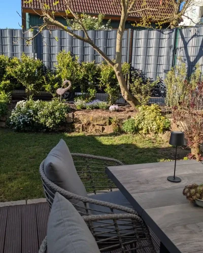 Nachher-Ansicht derselben Gartenfläche nach der Umgestaltung durch Molle GaLaBau: Moderne Holzterrasse, gemütliche Sitzgruppe, Natursteinmauer und blickdichter Sichtschutzzaun.